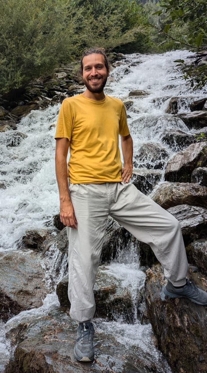 Una foto mía sonriendo y achinando los ojos porque el sol me pegaba de frente. Llevo puestos un sweater bordó y un jean que se ve apenas. Tengo una mano medio metida en el bolsillo porque nunca sé que hacer con las manos cuando me sacan fotos. De fondo se ven unos techitos de tejas rojas muy pintorescas